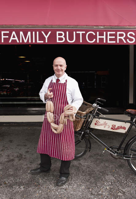 Açougueiro segurando salsicha na frente de sua loja — Fotografia de Stock