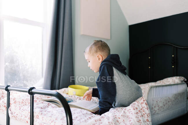 Kleiner Junge liest ein Buch auf dem Bett, um sich die Zeit zu vertreiben. — Stockfoto