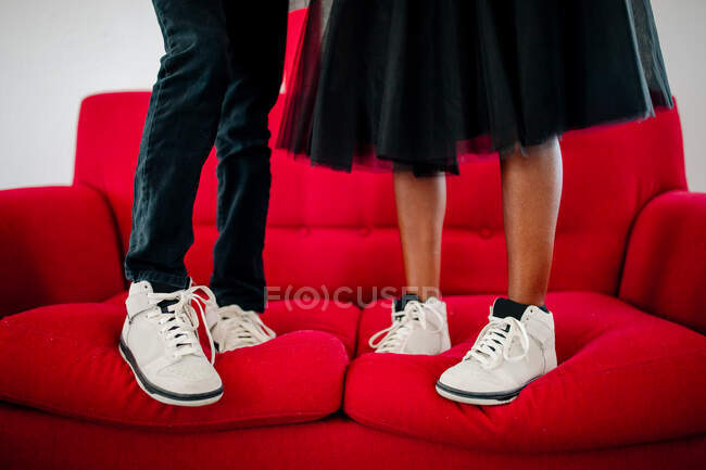 Нижние ноги мужчины и женщины в белых кроссовках стоят на красном диване — стоковое фото