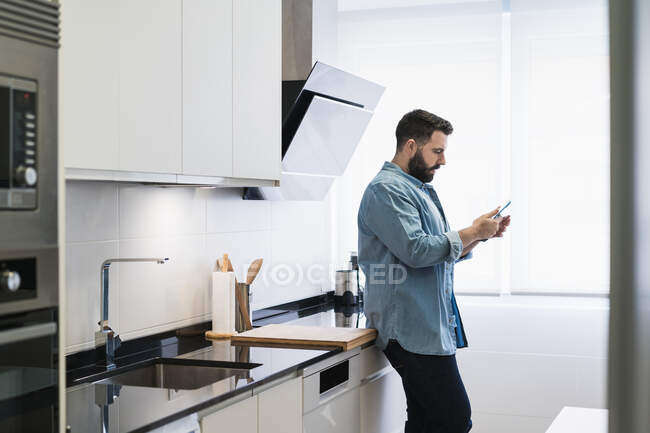 Homem com um telefone celular na cozinha em uma camisa jeans — Fotografia de Stock