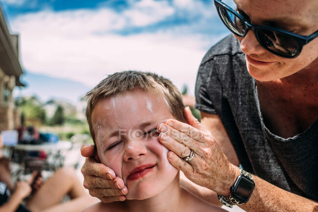 Nahaufnahme einer Mutter, die ihrem Sohn an einem Sommertag im Pool Lotion ins Gesicht sprüht — Stockfoto