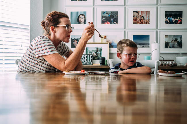 Мама предлагает кусочек торта плачущему сыну за кухонным столом — стоковое фото