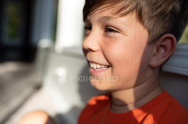 Sonriendo de cerca el perfil del niño sentado junto a la ventana interior - foto de stock