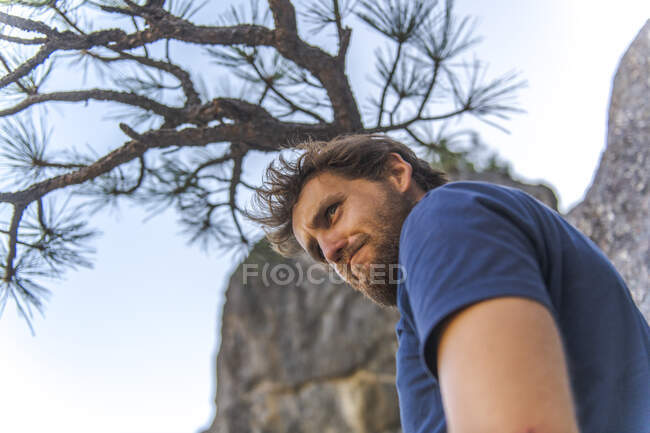 Mann im T-Shirt mit Bart macht lustiges Gesicht und schaut unter Baum hinunter — Stockfoto