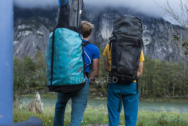 Два человека с большим рюкзаком стоят перед горой и рекой — стоковое фото