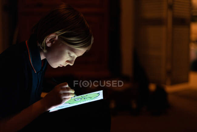 Profile of Tween drawing with stylus on tablet indoors low lit — Stock Photo