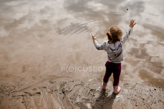 Toddler girl in rain boots throwing rock into lake in the summer — Stock Photo