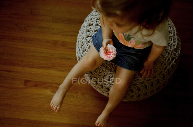 Jeune fille assise sur une bouffée tenant une fleur rose dans son salon — Photo de stock