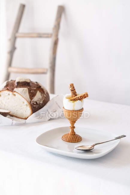 Dos galletas con semillas de sésamo en un huevo cocido roto en un soporte de madera sobre una mesa con un mantel blanco y pan en el fondo - foto de stock