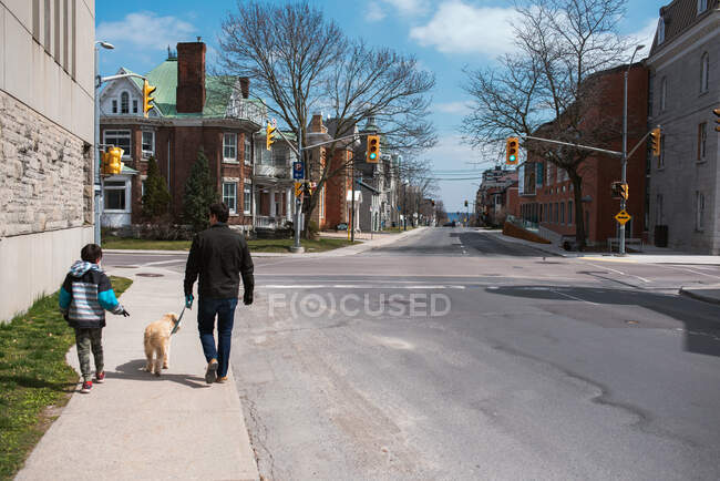 Padre e hijo paseando a un perro en la acera de una tranquila calle de la ciudad. - foto de stock