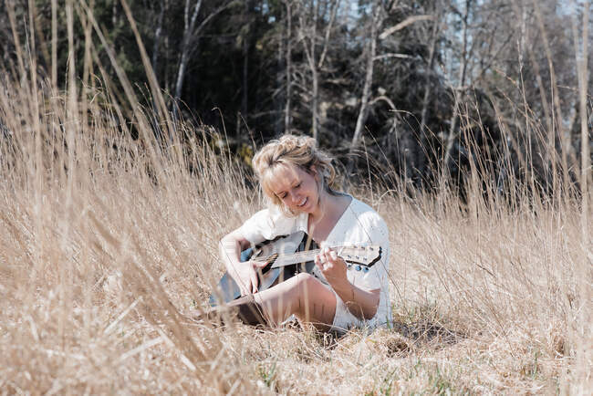 Donna seduto in un campo felicemente suonare la chitarra in estate — Foto stock