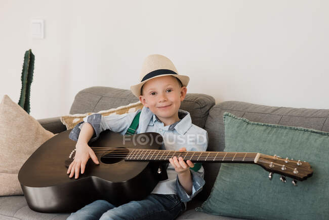 Хлопчик посміхається, граючи на гітарі з капелюхом вдома — стокове фото