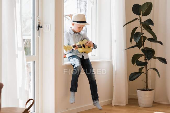 Young boy sat on a window ledge at home playing ukulele — Stock Photo