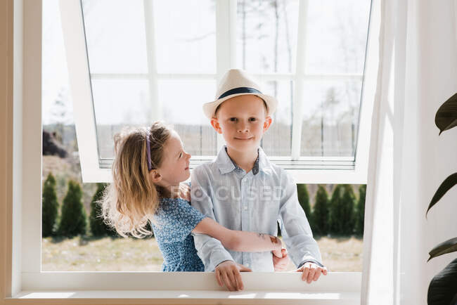 Brother and sister hugging whilst at home looking through a window — Stock Photo