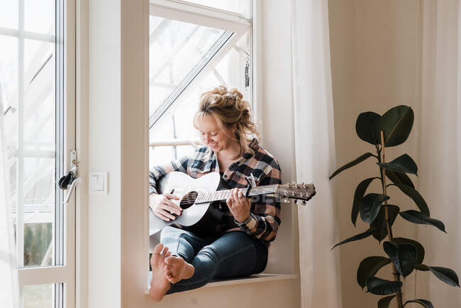 Woman sat at home on a window ledge smiling playing guitar — Stock Photo