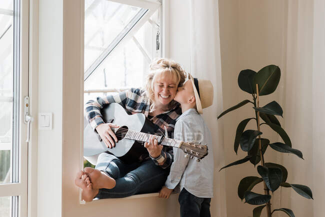 Woman sat playing guitar at home whilst her son gives her a kiss — Stock Photo