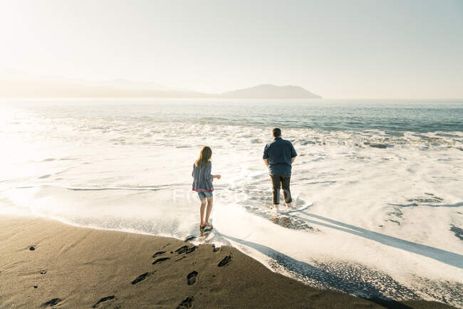 Pai e filha caminhando na água na praia durante o pôr do sol — Fotografia de Stock