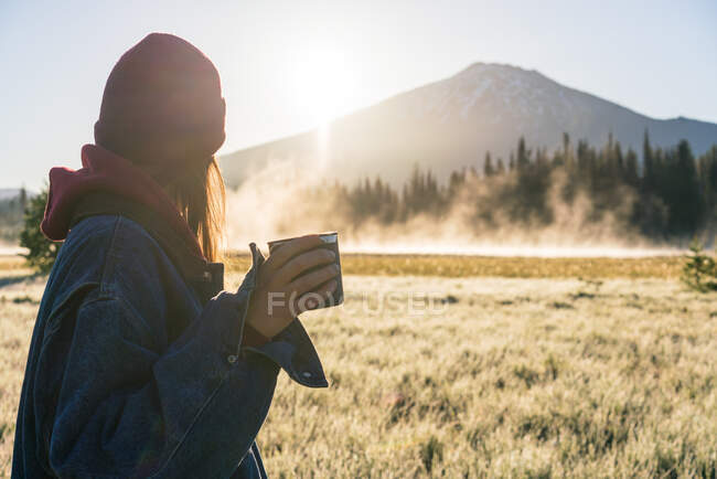 Молода жінка тримає каву на сході сонця, дивлячись на туманну гору — стокове фото