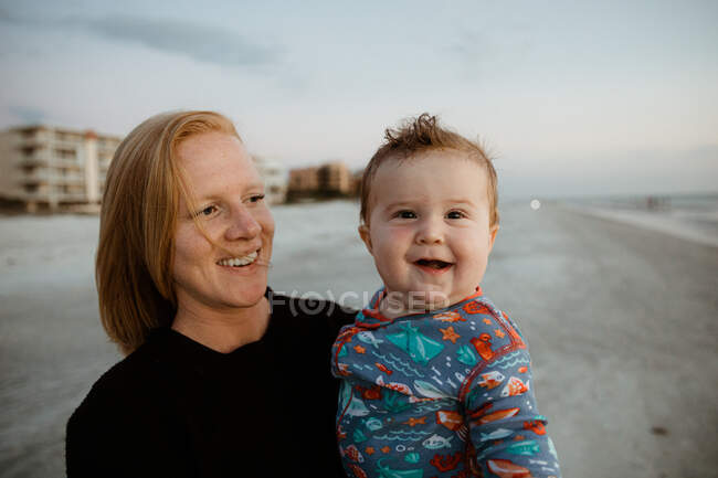 Толстый мальчик с кривой улыбкой, удерживаемый молодой рыжеволосой мамой на пляже — стоковое фото