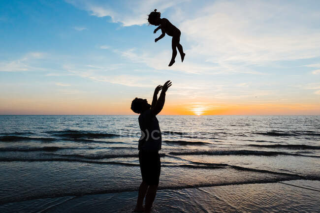 Pai joga sua filha no ar ao pôr do sol em férias na praia — Fotografia de Stock
