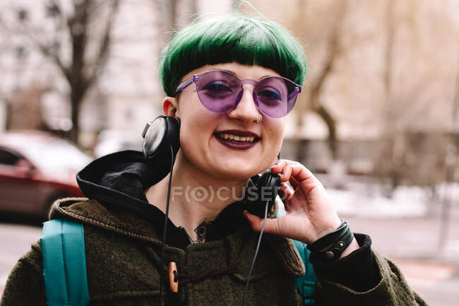 Retrato de hipster no binario feliz en gafas de sol púrpura de pie en la ciudad - foto de stock
