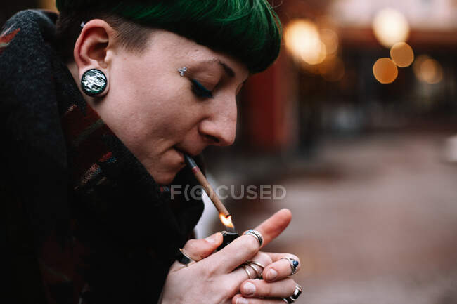 Portrait of non-binary hipster woman lighting a cigarette outdoors in winter — Stock Photo