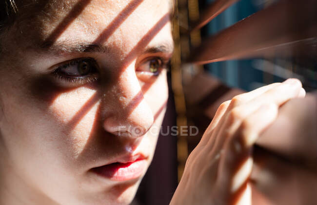 Chica joven con ojos verdes abriendo una cortina de madera con los dedos para mirar a través de la ventana mientras la luz del sol crea sol y sombra en su cara que muestra desesperanzado. Fotografía horizontal - foto de stock