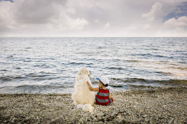 Toddler boy sitting with golden retriever dog on beach looking at lake — Stock Photo