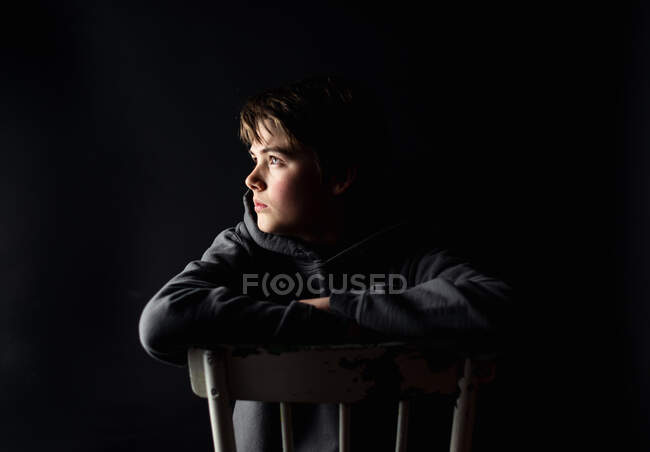 Низький ключовий портрет хлопчика-підлітка, що сидить на стільці в темній кімнаті . — стокове фото