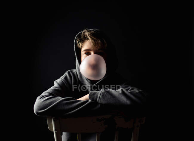 Хлопчик-підліток в балахон, сидить на стільці в темній кімнаті, дме бульбашку . — стокове фото