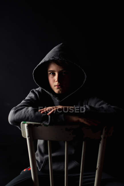 Портрет хлопчика-підлітка в светрі, що сидить на стільці в темній кімнаті . — стокове фото