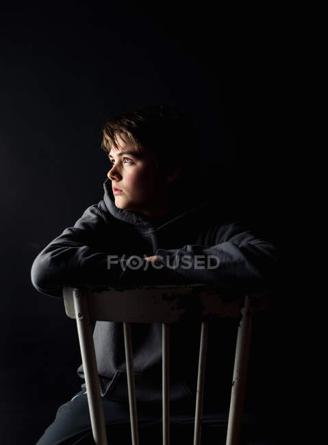 Baixo retrato chave do menino adolescente sentado em uma cadeira em um quarto escuro. — Fotografia de Stock