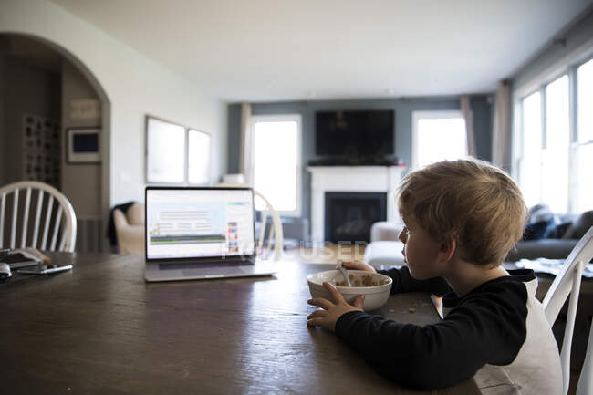 Vista lateral larga do menino loiro comendo aula de observação de cereais no laptop — Fotografia de Stock