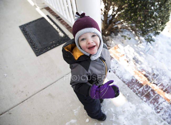 Niño en equipo de invierno de pie en el porche delantero nevado mira hacia arriba en la cámara - foto de stock