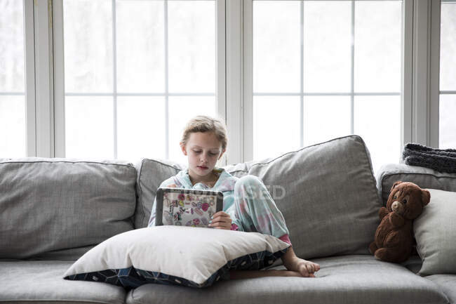 Blond mädchen zuhause krank aus schule plays tablet auf couch mit teddy — Stockfoto