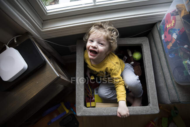 Laughing Boy se sienta en la caja de juguetes junto a los contenedores de almacenamiento mira hacia arriba en la cámara - foto de stock