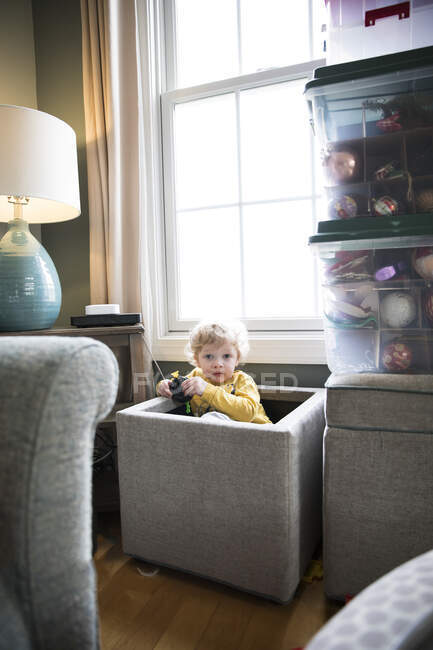 Câmera consciente criança senta-se em caixa de brinquedo na sala de estar na frente da janela — Fotografia de Stock