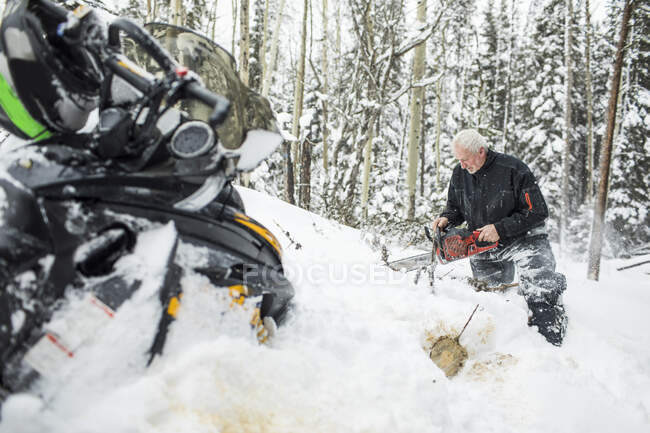 Retired elderly man using chainsaw to clear trails while snowmobiling. — Stock Photo