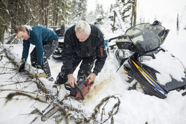 Elderly man and son using chainsaw to clear trails while snowmobiling. — Stock Photo