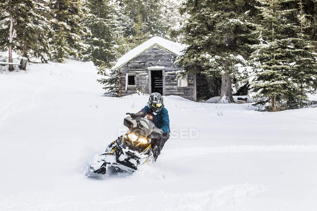 Confident man on snowmobile with old cabin behind. — Stock Photo