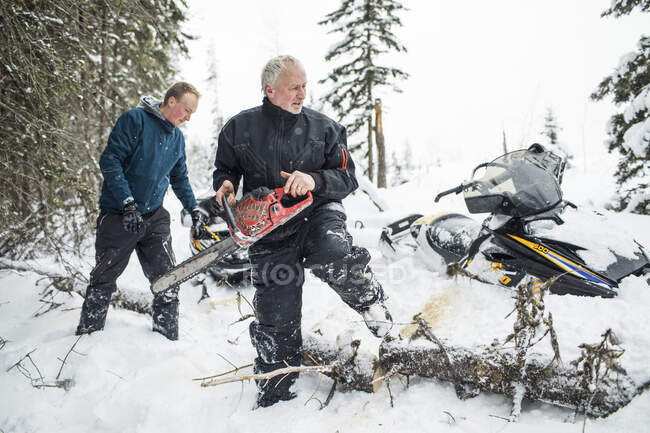 Retired man and his son using chainsaw to clear trails while snowmobiling — Stock Photo