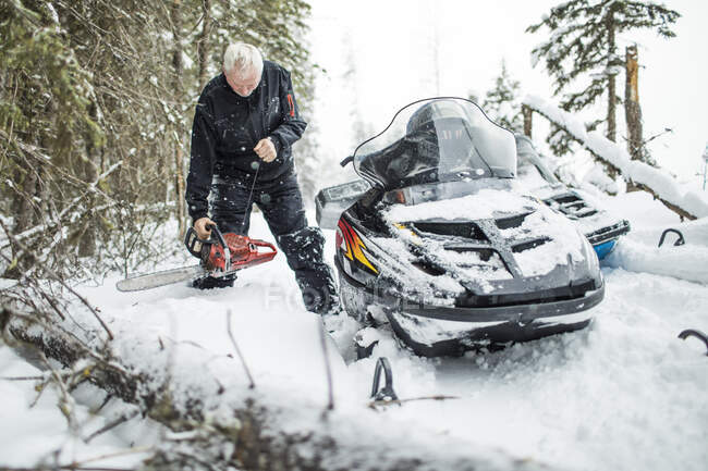 Retired man using chainsaw to clear trails while snowmobiling — Stock Photo