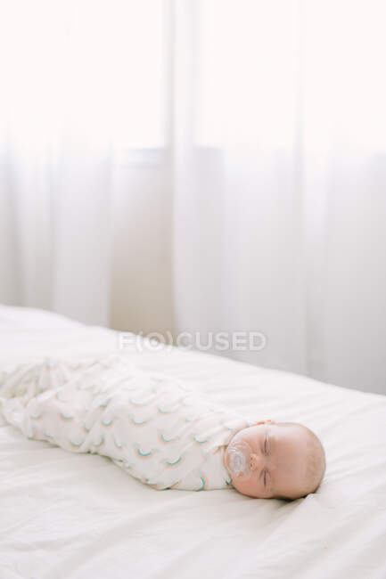 Nouveau-né dormant emmailloté dans une couverture arc-en-ciel sur lit blanc — Photo de stock