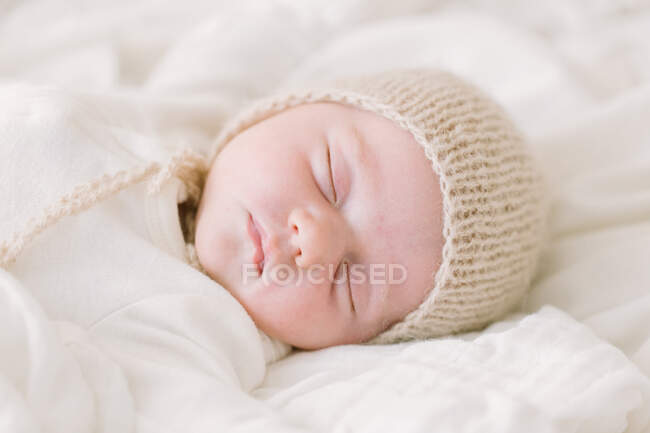 Newborn baby in knit bonnet sleeping on white bed — Stock Photo