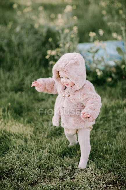 Baby girl first steps in pink fur coat outside on grass — Stock Photo