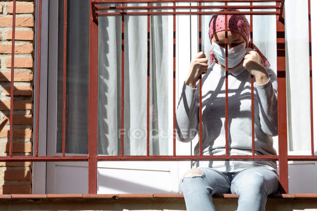 Una donna con cancro e depressione rinchiusa sul balcone. Foulard rosa e maschera viso. Coronavirus da quarantena. — Foto stock