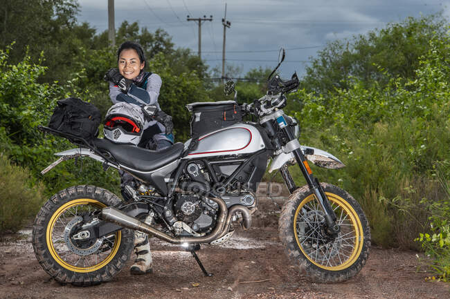 Mujer posando detrás de su motocicleta estilo scrambler en camino de tierra - foto de stock