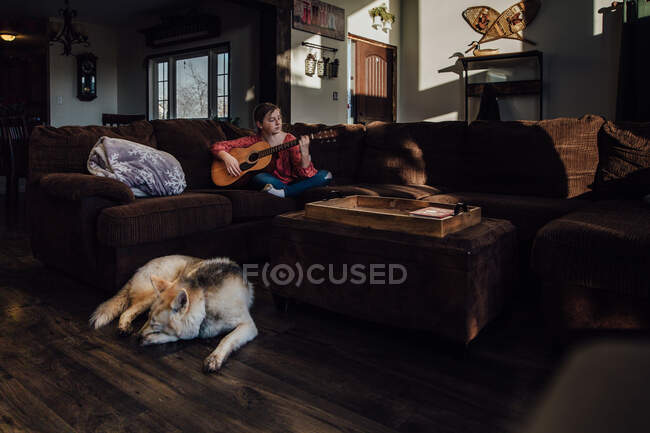 Молодая девушка играет на гитаре на диване с собакой спит поблизости — стоковое фото