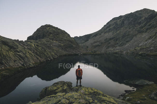 Un joven con mochila mirando el reflejo del lago en Sierra de Gredos, Ávila España - foto de stock