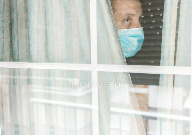Foto de un hombre blanco de mediana edad con mascarilla médica mirando por la ventana durante la cuarentena del coronavirus - foto de stock